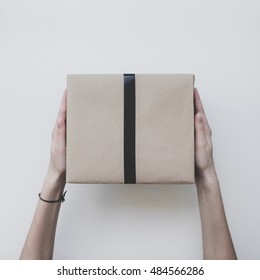 Parcel Wrapped In Kraft Paper With Black Ribbon, In Female Hands On A White Background. Minimalistic Photo. Womans Hand Holding Gift Box. Top View.