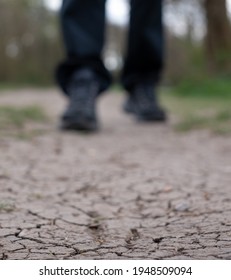 Parched earth photographed after a drought. Man walks along cracked mud path in the UK. Shoes and trouser bottoms only are visible.