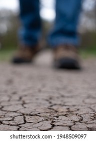 Parched earth photographed after a drought. Man walks along cracked mud path in the UK. Shoes and trouser bottoms only are visible.