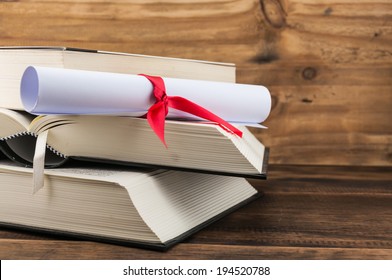 A Parchment Diploma Scroll, Rolled Up With Red Ribbon On Stack Of Book On Wood Background