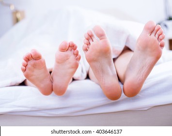 Parent And Child Feet In Bed Lolling Out From Under The Duvet.