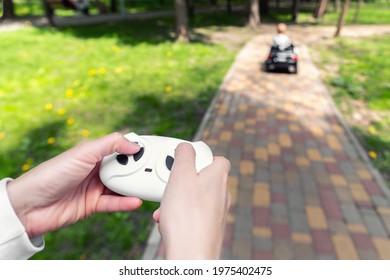 Parent Hand Holding Remote Controller Of Electric Powered Battery Car With Little Kid Boy Enjoy Having Fun Riding Toy Asphalt Path Road City Park At Summer Day. Mother Child Safety And Care Concept