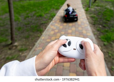 Parent Hand Holding Remote Controller Of Electric Powered Battery Car With Little Kid Boy Enjoy Having Fun Riding Toy Asphalt Path Road City Park At Summer Day. Mother Child Safety And Care Concept