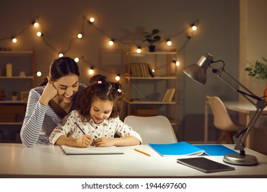Parent helping child. Happy family doing homework in the evening. Mother and daughter working on school assignment sitting at desk with lamp in cozy dark room with LED lights. Kids learning concept - Shutterstock ID 1944697600
