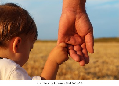 A parent holds the hand of a lovely child. Father and son holding hands in the background of the yellow field of wheat and blue sky. - Powered by Shutterstock