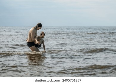 A parent lifts a child in the ocean, both wearing swimwear. The sky is overcast, and the water stretches to the horizon - Powered by Shutterstock