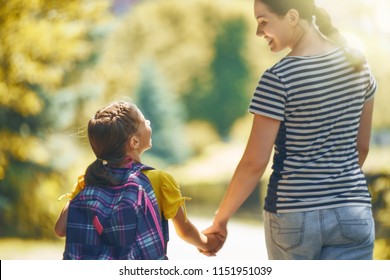 Parent And Pupil Of Primary School Go Hand In Hand. Woman And Girl With Backpack Behind The Back. Beginning Of Lessons. First Day Of Fall.