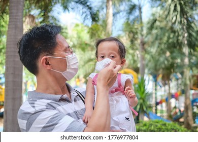 Parent Putting Mask On His Asian Toddler Girl Child, Father And Daughter Wearing Protective Medical Mask In Public Playground Area During The Covid-19 Health Crisis, New Normal Lifestyle Concept