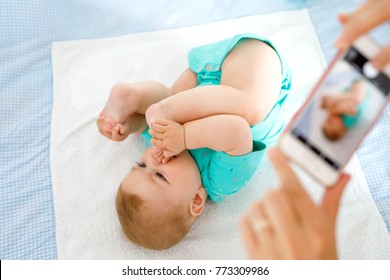 Parent Taking Photo Of A Baby With Smartphone. Adorable Newborn Child Taking Foot In Mouth. Sucking Feet. Digital Family Memories