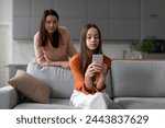 Parental monitoring of teens. Curious mother spying daughter, girl messaging on smartphone on sofa at home. Family and modern technologies