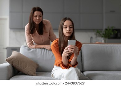 Parental monitoring of teens. Curious mother spying daughter, girl messaging on smartphone on sofa at home. Family and modern technologies