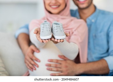 Parenthood concept. Cute baby shoes in pregnant muslim couple hands. Unrecognizable arab man and woman showing small shoes for their coming baby, closeup