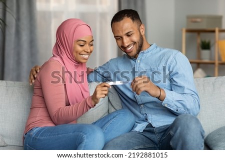 Parenthood Concept. Happy Black Muslim Spouses Holding Positive Pregnancy Test While Sitting On Couch At Home, Smiling Islamic Couple Getting Ready For Childbirth, Celebrating Good News