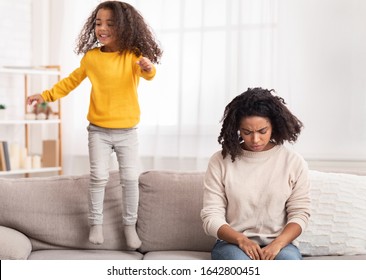 Parenting Hyperactive Kid. Exhausted Black Mother Sleeping While Daughter Jumping On Sofa At Home