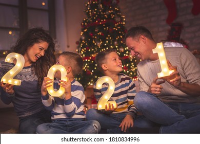 Parents Celebrating New Years Eve At Home With Kids, Sitting By The Christmas Tree, Holding Illuminative Numbers 2021 Representing The Upcoming New Year