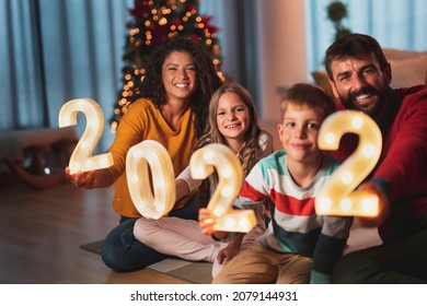 Parents Celebrating New Years Eve At Home With Kids, Sitting By The Christmas Tree, Holding Illuminative Numbers 2022 Representing The Upcoming New Year