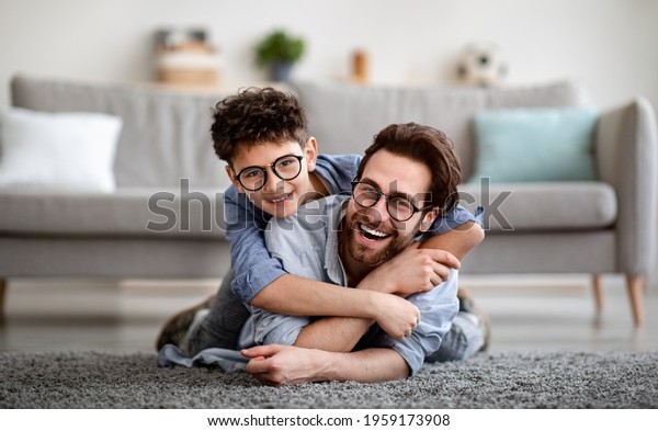Parents and children being friends. Joyful father\
and son having fun, dad lying on floor, carrying boy on back and\
smiling together to camera, spending tim at home together. Single\
dad concept