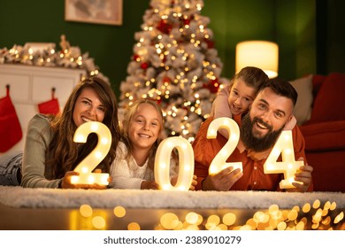 Parents and children having fun celebrating New Year together at home, lying by the Christmas tree holding illuminative numbers 2024 - Powered by Shutterstock