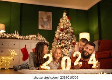 Parents and children having fun celebrating New Year together at home, lying by the Christmas tree holding illuminative numbers 2024 - Powered by Shutterstock