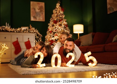 Parents and children having fun celebrating New Year together at home, lying by the Christmas tree holding illuminative numbers 2025 - Powered by Shutterstock