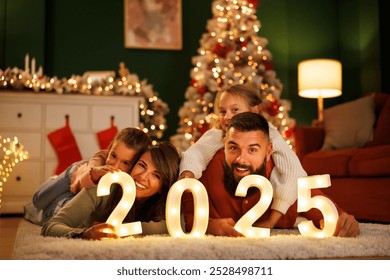 Parents and children having fun celebrating New Year together at home, lying by the Christmas tree holding illuminative numbers 2025 - Powered by Shutterstock