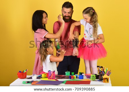 Parents and children paint on fathers arms with gouache. Family leisure time and art concept. Girls, man and woman with happy faces by art desk on yellow background. Artists put paint on guys body