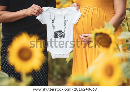 Parents expecting a baby hold a photography - themed onesie in a sunflower field in Maryland.