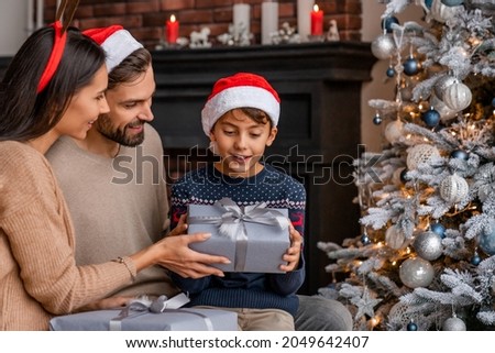 Parents giving present their son while sitting near Christmas tree in living room. Little boy opening Christmas present, Happy New Year and Merry Christmas