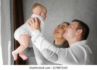 Parents Hold An Infant At Arms Length. Parents With An Infant. Smiling Mother And Father Holding Their Newborn Baby Daughter At Home