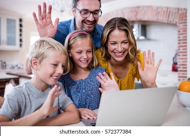 Parents And Kids Waving Hands While Using Laptop For Video Chat In Kitchen