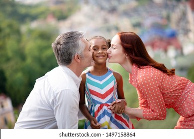 Parents kissing. Adoptive parents kissing their cute dark-skinned girl while walking in the park