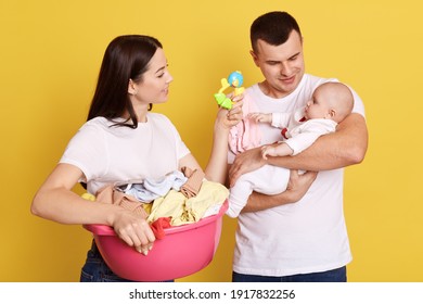 Parents looking after newborn child while doing household chores and laundry, mother with dark hair showing beanbag to their little kid, fatter holds baby in hands, isolated over yellow background.