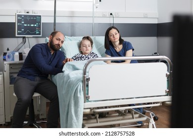 Parents and sick little girl watching television comedy program while sitting in pediatric clinic ward recovery room. Family spending time together in hospital patient saloon while enjoying tv show.