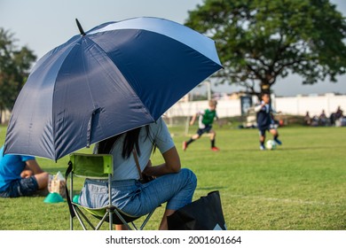 Parents Sitting And Watching Their Son Playing Football In A School Tournament On A Clear Sky And Sunny Day. Sport, Outdoor Activity With Lifestyle And A Happy Family. Soccer Mom And Dad Concept.