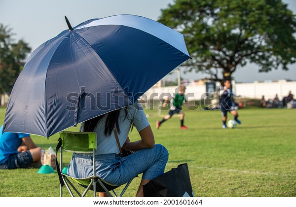 Parents sitting and watching their son playing\
football in a school tournament on a clear sky and sunny day.\
Sport, outdoor activity with lifestyle and a happy family. Soccer\
mom and dad concept.