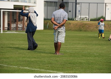 Parents Standing And Watching Their Son Playing Football In A School Tournament On A Sideline With A Sunny Day. Sport, Outdoor Active, Lifestyle, Happy Family And Soccer Mom And Soccer Dad Concepts.