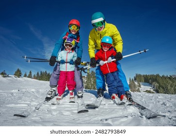 Parents teach kids to ski hold between legs wearing bright skiing outfits and protective helmets, are captured smiling on a sunlit slope with a forest in the background