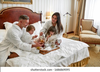 Parents And Two Kids In White Bathrobes Smiling While Having Breakfast In Bed, Eating And Drinking In Luxurious Hotel Room. Family, Resort, Room Service Concept. Horizontal Shot