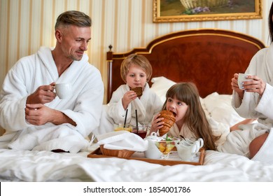 Parents And Two Kids In White Bathrobes Having Breakfast In Bed, Eating Pastries And Drinking Coffee In Luxurious Hotel Room. Family, Resort, Room Service Concept. Horizontal Shot