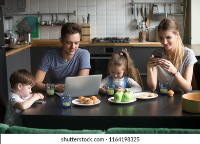 Parents using laptop and smartphones having breakfast with children, smiling dad reading morning online computer news while mom and kids entertaining with phones apps, family gadget addiction concept - Powered by Shutterstock