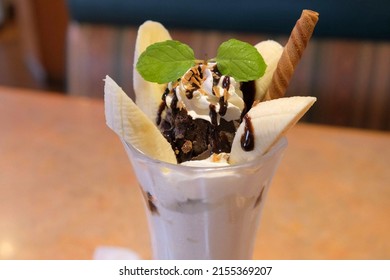 A Parfait Topped With Banana, Chocolate Brownie, Banana, Whipped Cream, Herbs, Crunch And Chocolate Syrup On Top Of Ice Cream