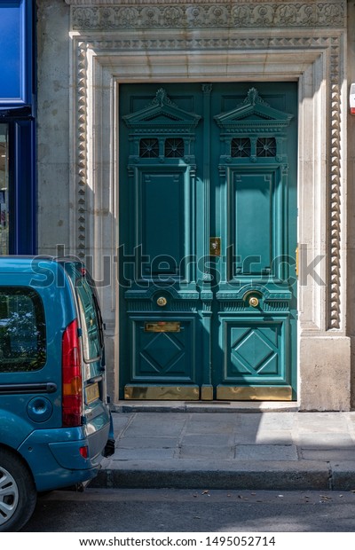 Paris / France - 06 26 2019: Green door of antique\
building with golden shiny doorknobs and details. Vintage ornate\
wooden door painted in sea-green. Blue car at European city street\
in front of door