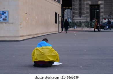 Paris, France - April 2022 - A man, wrapped in a yellow and blue Ukrainian flag, sits alone on the sidewalk, in the Louvre neighborhood, likely to draw attention on the war between Ukraine and Russia