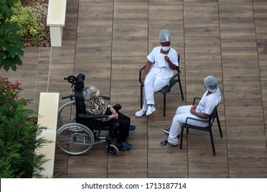 PARIS, FRANCE - APRIL 24, 2020: Nurses Caring For Elderly Or Disabled People In A Nursing Home During The Coronavirus Pandemic Covid-19. Carers Having A Break In Th Sun With An Old Woman Resident.