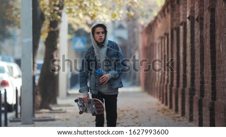 Paris, France - April 5, 2018: Unrecognizable young rebellious teenage boy in jeans wear and balaclava walks with skateboard and can spray of paint down the street, stops to paint the street wall