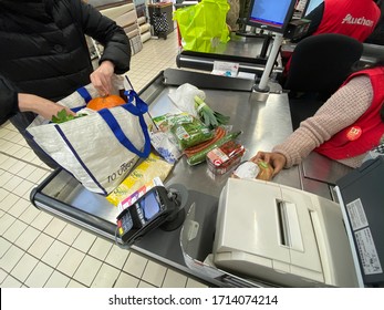 Paris, France - Dec 21, 2020: Overhead View Of Woman At Packing In Her Bag Multiple Products Bought French Auchan Supermarket Customers Silhouettes In Background - Shopping Buying Consumerism Concept