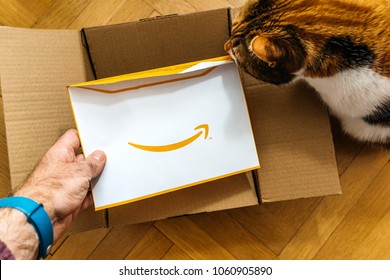 PARIS, FRANCE - FEB 14, 2018: Man unboxing Amazon.com cardboard containing a surprise yellow envelope with Greeting Card and large smiling Amazon logotype from friend Amazon being helped by cat
