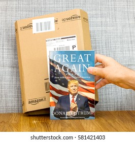 PARIS, FRANCE - FEB 9, 2017: Man Holding Book Great Again - How To Fix Our Crippled America Biography Book By Donald J Trump In Front Of Amazon Delivery Box. Crippled America Is A Book By Donald Trump