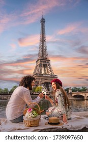 PARIS, FRANCE - Jun 07, 2019: A Happy Smiling Couple Toasts Their Love With Wine And Flowers On A Picnic Set. In Front Of Them The Eiffel Tower And The Seine River At Sunset