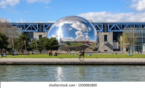 PARIS, FRANCE - MARCH 19, 2019: People Enjoying The Sunny Springtime In The Parc De La Villette. A Man Is Performing A Wheelie Stunt With His Bike In Front La Géode Dome, Along The Canal De L'Ourcq. 